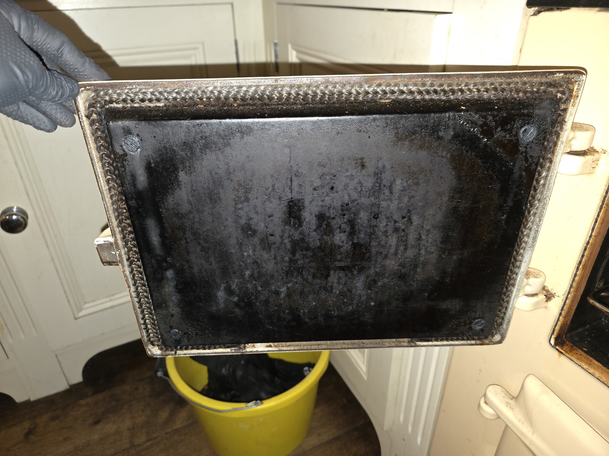 A gloved hand holding a heavily burnt oven tray before cleaning
